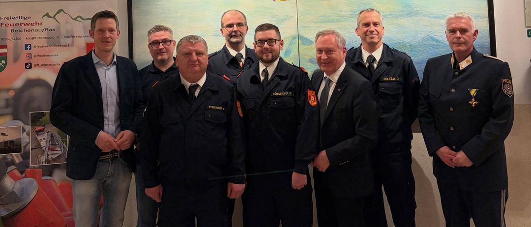 Mehrere Männer in Uniformen stehen zusammen und lächeln für ein Foto. Sie stehen vor einem großen Banner mit deutschem Text und einer Illustration eines Feuerwehrwagens.