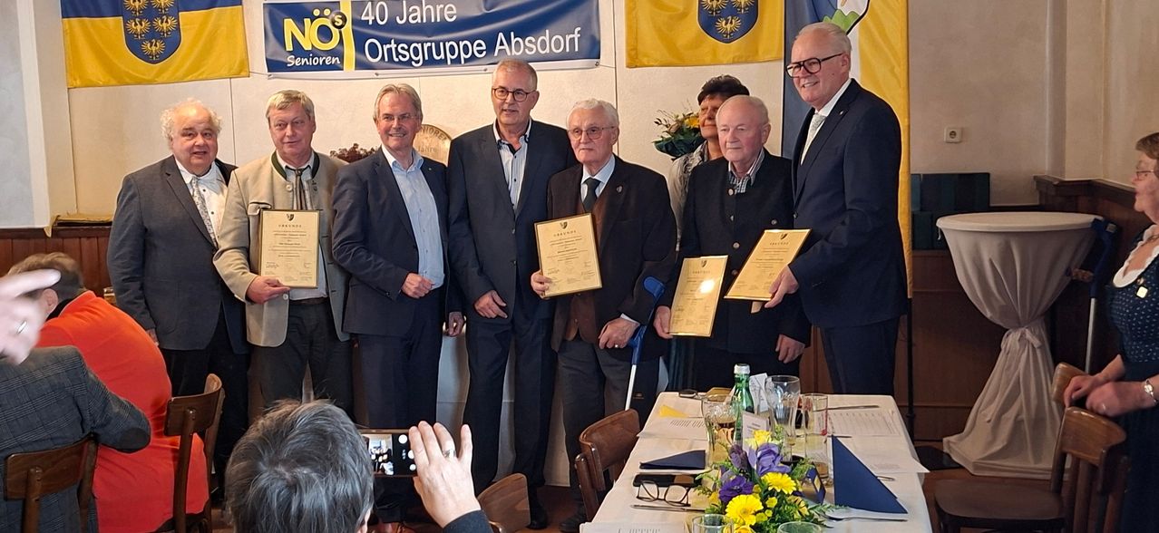 Eine Gruppe von Männern in Anzügen steht mit Urkunden in der Hand und lächelt für ein Foto bei einer Veranstaltung. Ein Banner steht '40 Jahre Ortsgruppe Absdorf', und es gibt Flaggen und Blumen im Hintergrund.