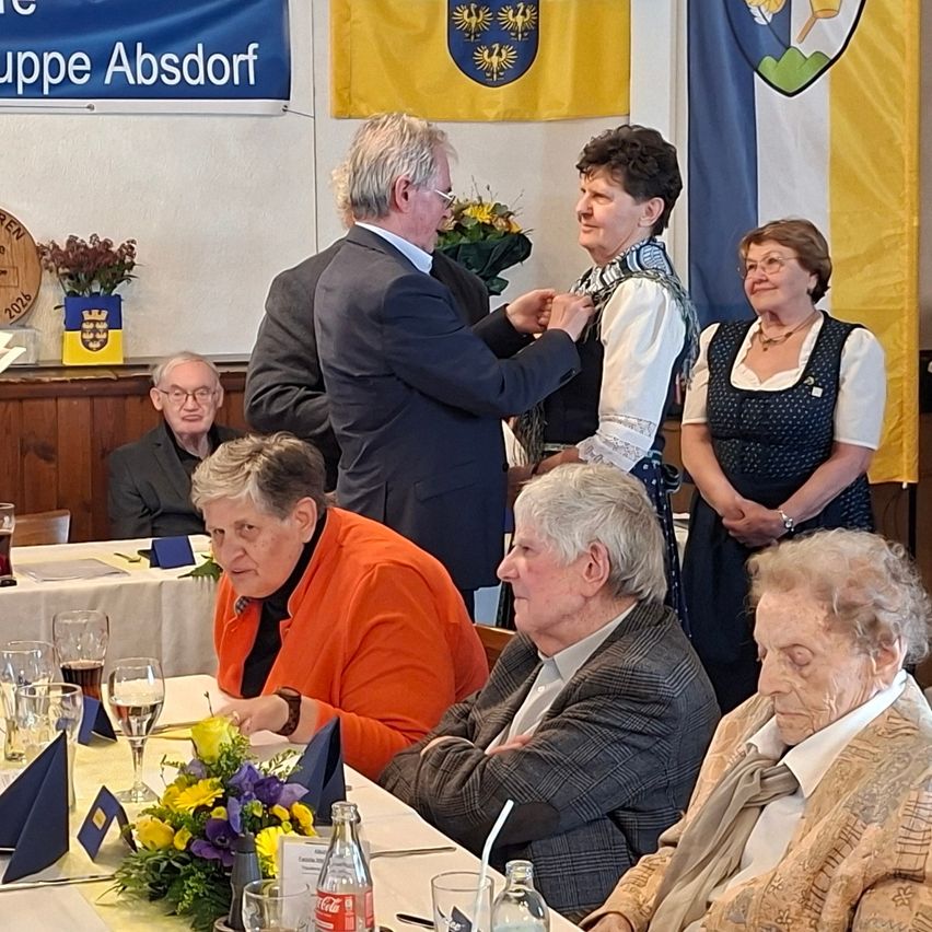 Mehrere ältere Menschen versammeln sich an einem Tisch. Ein Mann in Anzug steht und scheint einer Frau eine Medaille zu geben. Zwei weitere Frauen stehen hinter ihnen. Es hängen Flaggen an der Wand hinter ihnen.