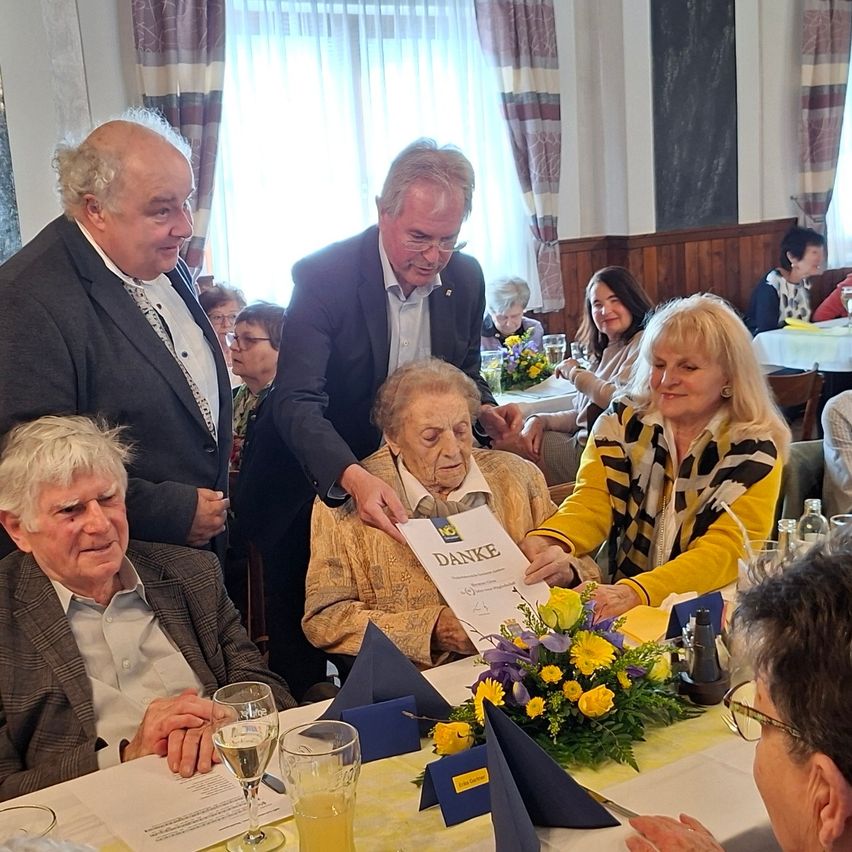 Eine Gruppe von Menschen, darunter eine ältere Frau, versammelt sich in einem Restaurant. Zwei Männer halten eine Urkunde und präsentieren sie der älteren Frau. Eine Frau mit einem Schal lächelt und schaut die ältere Frau an. Ein Tisch mit Blumen, Gläsern und einer Flasche steht vor ihnen. Hinter ihnen sitzen andere Menschen an einem Tisch mit Blumen und Weingläsern.