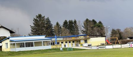 A yellow building with blue trim stands in a large grassy field. It has a blue umbrella outside and a sign reading 'SV Schutzen'. There are trees in the background and a cloudy sky.