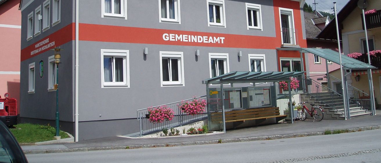 Ein graues Gebäude mit rotem Dach und weißen Zierleisten hat das Wort Gemeindeamt auf der Vorderseite. Eine Bushaltestelle mit einer Bank und Blumen steht davor.