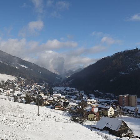 Ein verschneites Dorf umgeben von Bergen unter einem blauen Himmel mit Wolken. Mehrere Gebäude sind schneebedeckt.