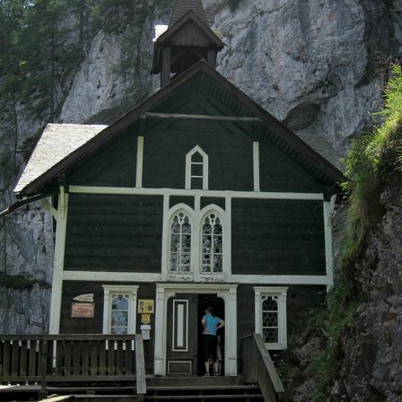 Eine kleine Holzkirche mit einem Glockenturm, in einen Felsen eingebettet. Eine Person steht in der Tür, umgeben von Grün.
