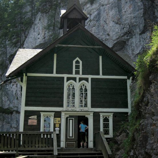 Eine kleine Holzkirche mit einem Glockenturm, in einen Felsen eingebettet. Eine Person steht in der Tür, umgeben von Grün.