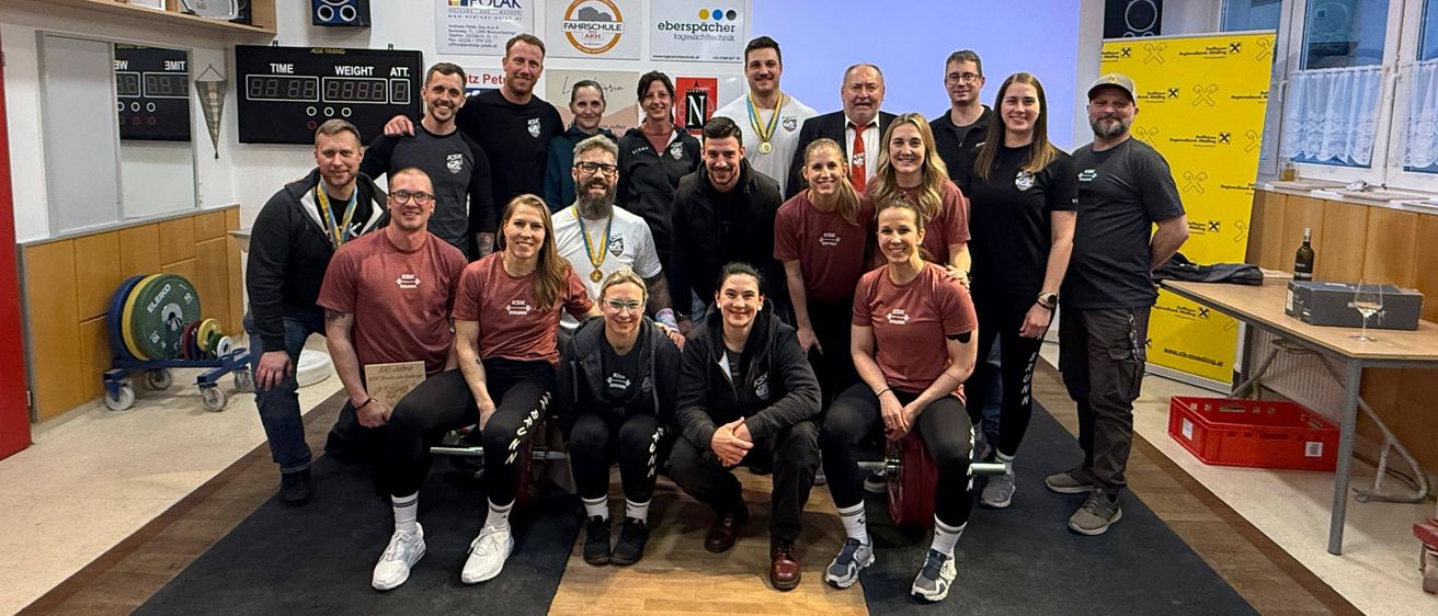 Eine Gruppe von Menschen mit Medaillen posiert für ein Foto in einem Fitnessstudio mit Holzboden und gelber Wand.