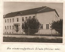 Schwarz-Weiß-Foto einer Schule mit der Bildunterschrift 'Die wiederaufgebaute Schule (Oktober 1950)'. Das Gebäude hat ein Satteldach, viele Fenster und ist von einem Zaun umgeben.