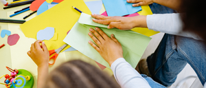 Two people are sitting and making paper crafts. A person is holding a pair of scissors. There is a pencil on top of the yellow paper.