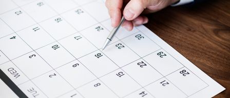 A hand holding a pen points at a calendar with dates marked, likely for planning or scheduling.