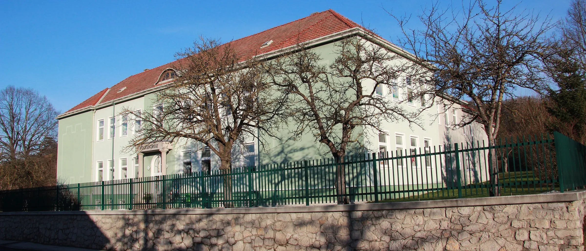Ein Gebäude mit rotem Dach und grünem Zaun ist an einem sonnigen Tag. Es hat viele Fenster und zwei kahle Bäume stehen davor.