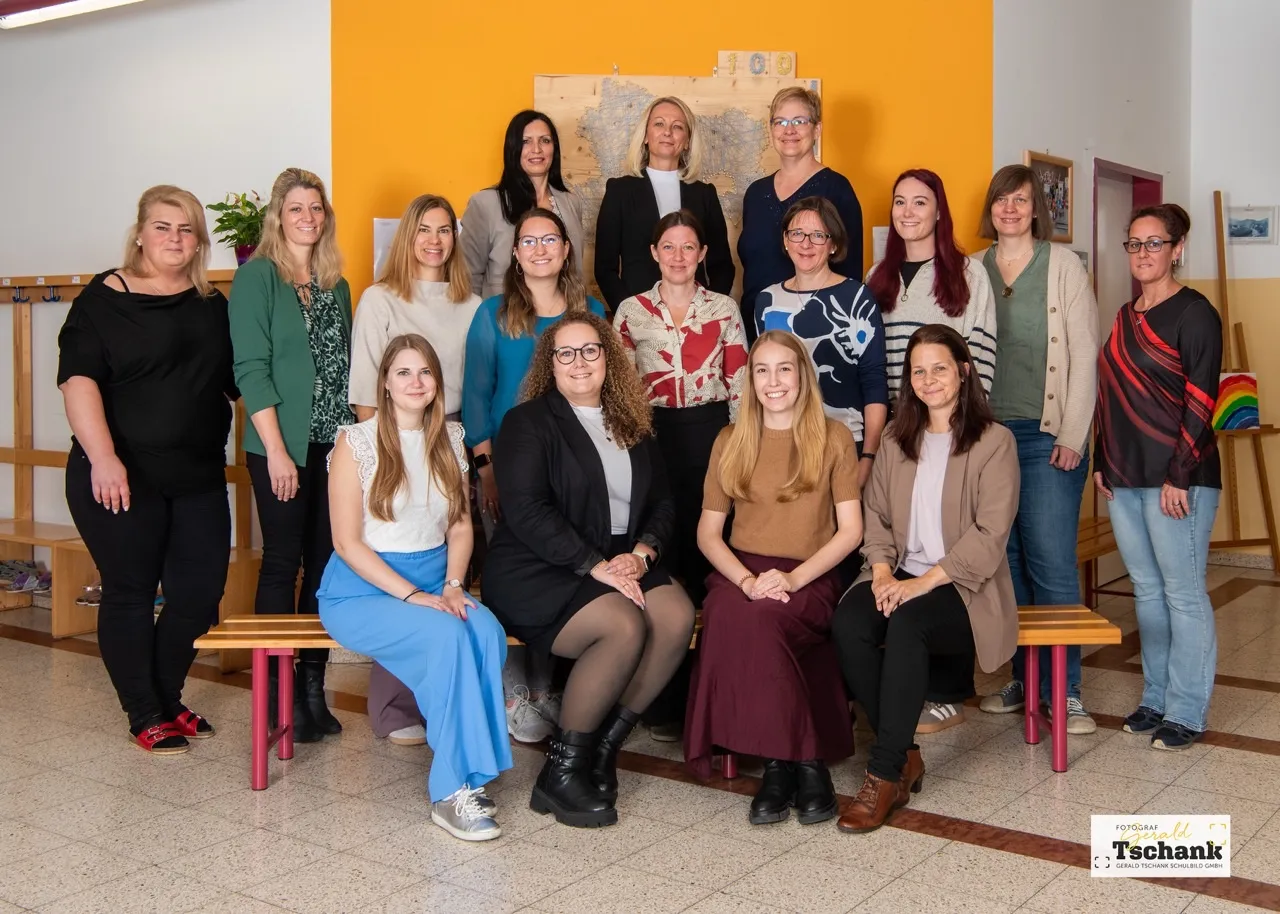 Eine Gruppe von Frauen, möglicherweise ein Team, posiert für ein Foto in einem Raum mit gelben Wänden. Sie sitzen auf Bänken und stehen dahinter. Eine Karte ist an der Wand hinter ihnen angebracht.