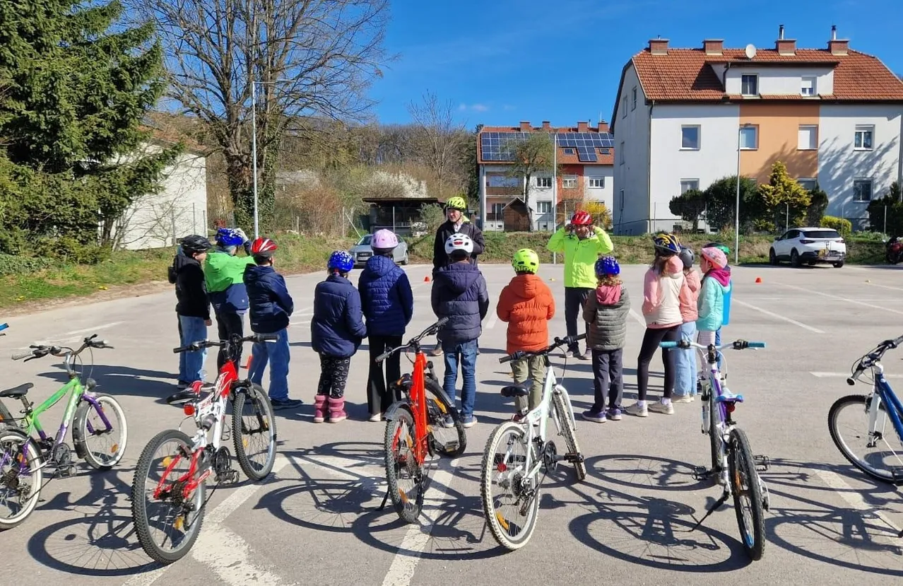 Eine Gruppe von Kindern und Erwachsenen auf einem Parkplatz mit Fahrrädern, die Helme und teilweise Jacken tragen, unter einem blauen Himmel.