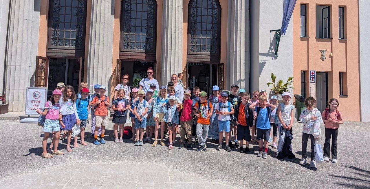 Eine Gruppe von Kindern und Erwachsenen steht vor einem Gebäude mit Bogenfenstern. Einige tragen Hüte und haben Rucksäcke dabei.