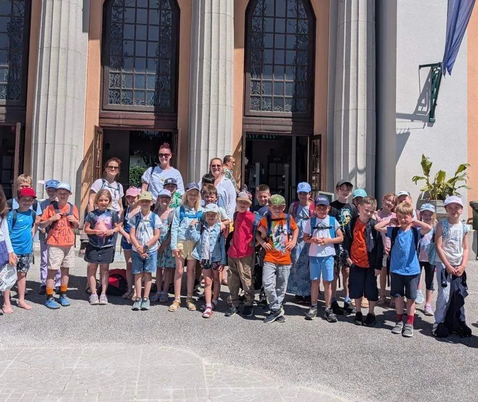 Eine Gruppe von Kindern und Erwachsenen steht vor einem Gebäude mit Bogenfenstern. Einige tragen Hüte und haben Rucksäcke dabei.