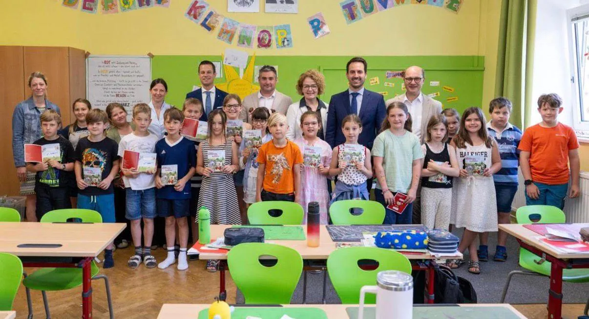 Eine Gruppe von Kindern und Erwachsenen steht in einem Klassenzimmer, hält Bücher und lächelt. Dahinter stehen zwei Erwachsene in der Nähe eines Tisches mit einem Thermos, einer Tasche und anderen Gegenständen. Das Klassenzimmer ist mit farbenfrohen Dekorationen und einer Tafel ausgestattet.