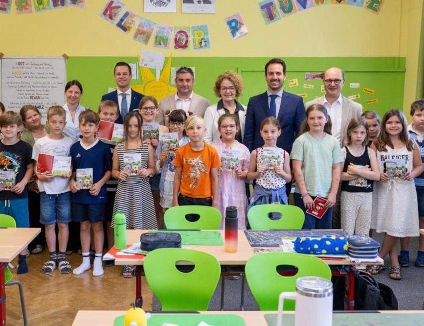 Eine Gruppe von Kindern und Erwachsenen steht in einem Klassenzimmer, hält Bücher und lächelt. Dahinter stehen zwei Erwachsene in der Nähe eines Tisches mit einem Thermos, einer Tasche und anderen Gegenständen. Das Klassenzimmer ist mit farbenfrohen Dekorationen und einer Tafel ausgestattet.