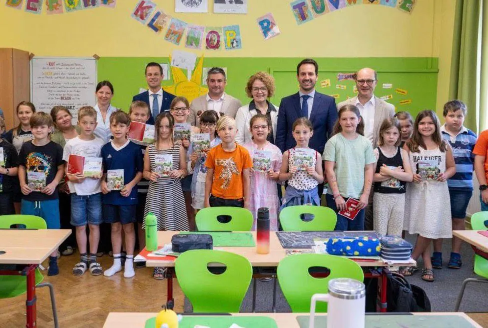 Eine Gruppe von Kindern und Erwachsenen steht in einem Klassenzimmer, hält Bücher und lächelt. Dahinter stehen zwei Erwachsene in der Nähe eines Tisches mit einem Thermos, einer Tasche und anderen Gegenständen. Das Klassenzimmer ist mit farbenfrohen Dekorationen und einer Tafel ausgestattet.