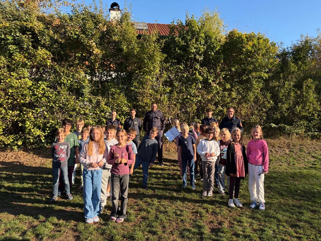 Eine Gruppe von Kindern und Erwachsenen steht in einer Reihe auf einer Grasfläche, mit Bäumen und einem Haus im Hintergrund.