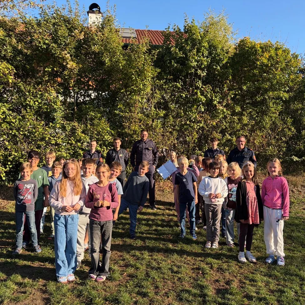 Eine Gruppe von Kindern und Erwachsenen steht in einer Reihe auf einer Grasfläche, mit Bäumen und einem Haus im Hintergrund.