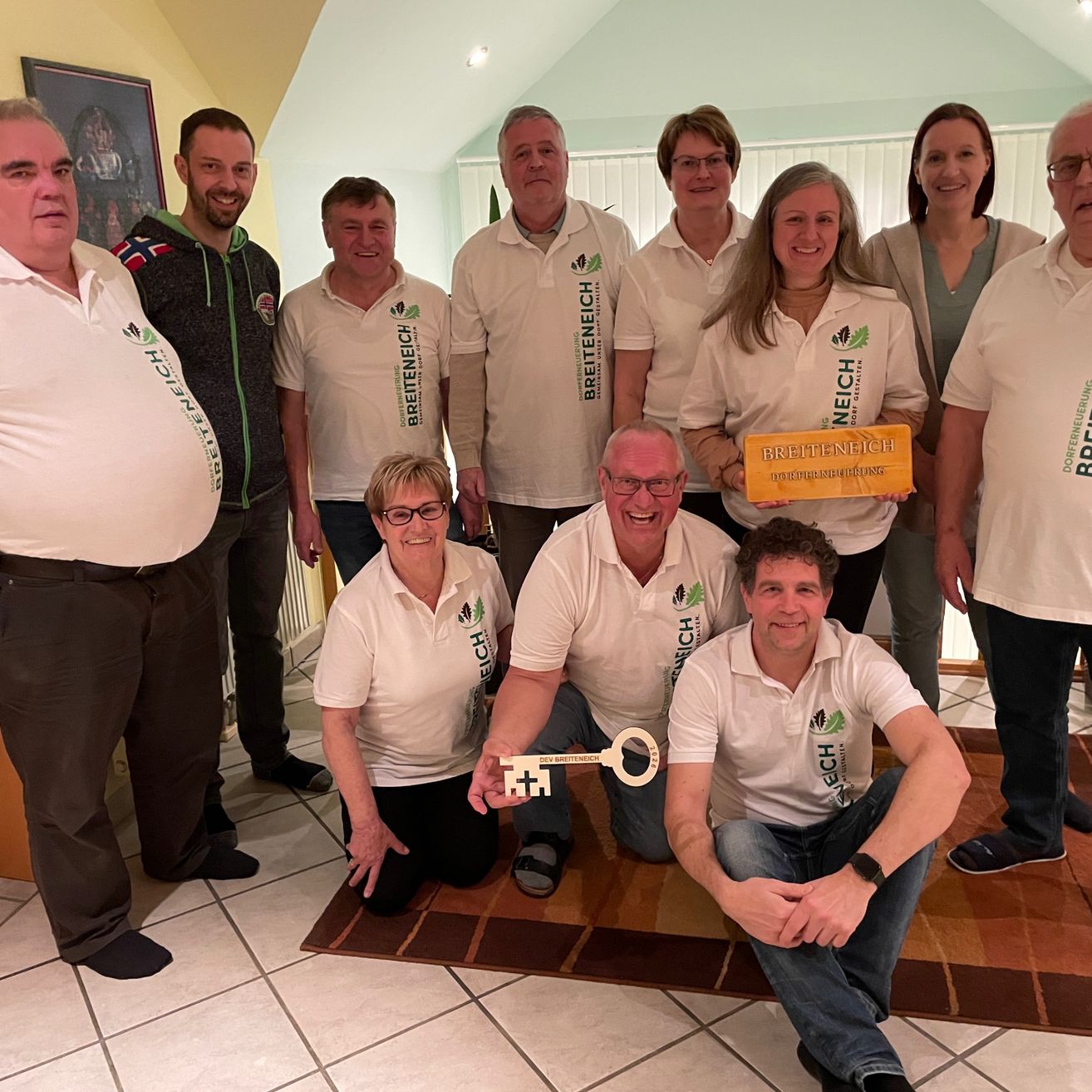 Eine Gruppe von Personen in weißen Hemden versammelt sich drinnen, lächelt und posiert für ein Foto. Zwei Männer halten einen Schlüssel und ein gelbes Schild.