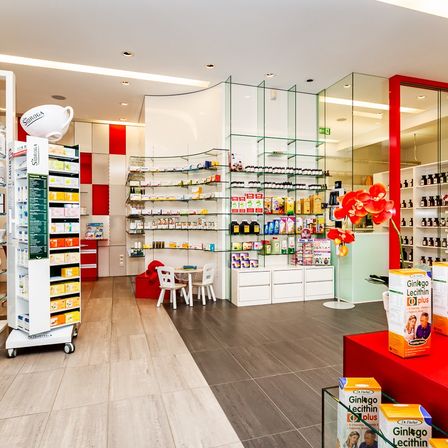Eine Innenapotheke mit Regalen voller Produkte. Ein Tisch und Stühle sind neben den Regalen angeordnet. Eine rote Blume steht auf einer Theke. Kartons von 'Ginko Lecithin' sind auf einem roten Tisch. Eine rot-weiß karierte Wand ist auf der linken Seite.