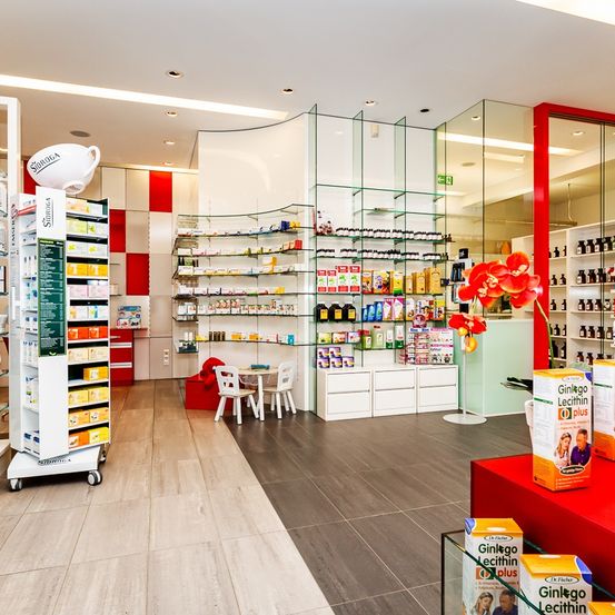 Eine Innenapotheke mit Regalen voller Produkte. Ein Tisch und Stühle sind neben den Regalen angeordnet. Eine rote Blume steht auf einer Theke. Kartons von 'Ginko Lecithin' sind auf einem roten Tisch. Eine rot-weiß karierte Wand ist auf der linken Seite.