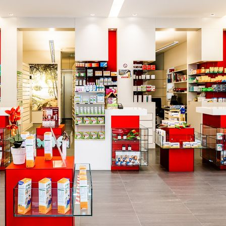 Eine Apotheke mit weißen und roten Wänden, ausgestattet mit Regalen, Produkten, Glasdisplays und einem roten und weißen Tresen. Pflanzgefäße und Blumen verleihen einen Hauch von Natur.
