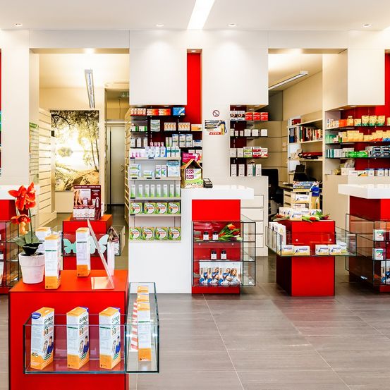 Eine Apotheke mit weißen und roten Wänden, ausgestattet mit Regalen, Produkten, Glasdisplays und einem roten und weißen Tresen. Pflanzgefäße und Blumen verleihen einen Hauch von Natur.