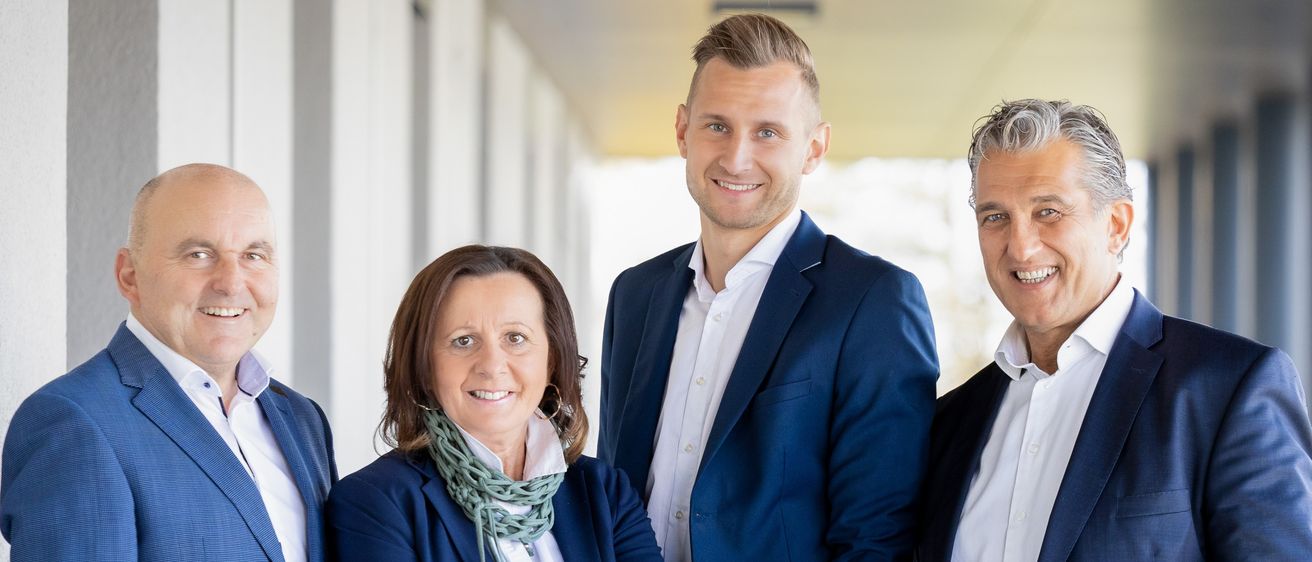 Vier Menschen in Anzügen stehen in einem Korridor und lächeln für ein Foto. Die beiden Männer an den Seiten tragen dunkle Anzüge, während die Frau in der Mitte einen blauen Blazer und eine Schürze trägt. Der Mann in der Mitte hat eine Uhr am Handgelenk.