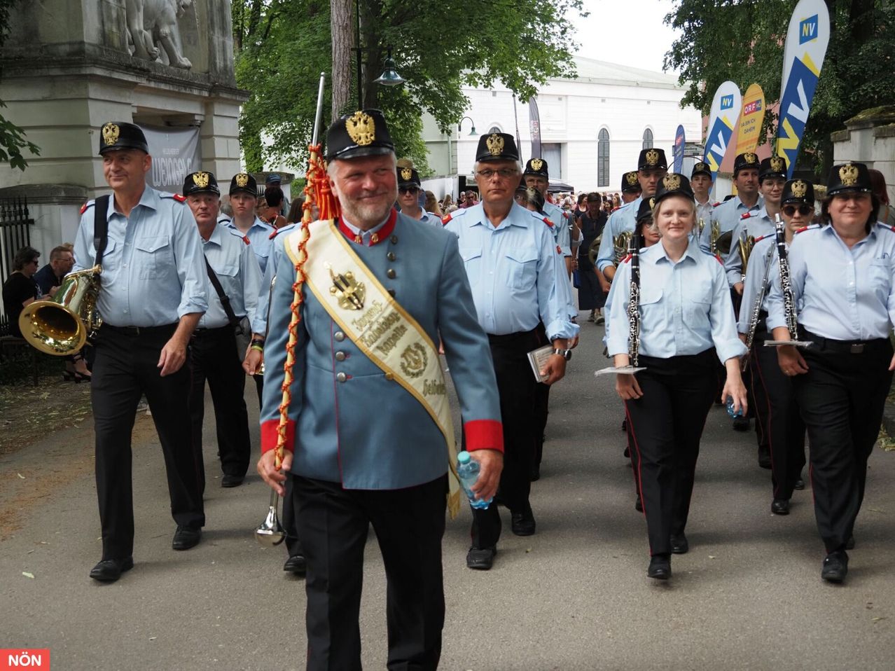 Eine Marschkapelle tritt bei einer Parade auf. Sie sind in Uniformen gekleidet und halten Musikinstrumente. Der Mann in der Mitte trägt eine Schärpe. Hinter ihnen stehen Bäume und ein Gebäude.