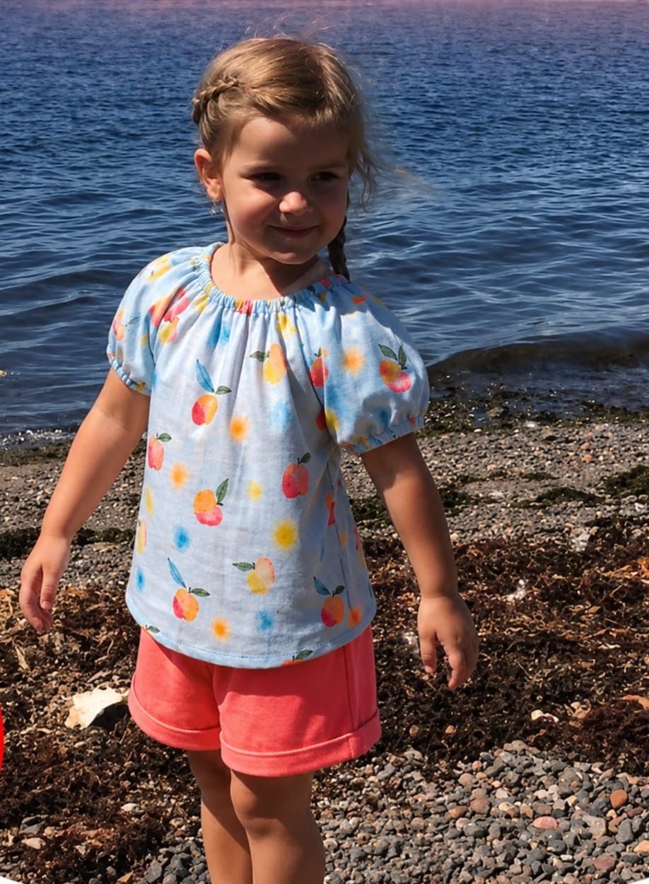 Ein kleines Mädchen in pfirsichfarbener Shorts und einem Top mit Pfirsich-Aufdruck steht am Ufer in der Nähe des Wassers. Sie schaut mit einem glücklichen Ausdruck nach vorne.