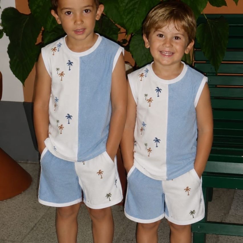 Zwei junge Jungen stehen nebeneinander und lächeln. Sie tragen passende blaue und weiße Outfits mit Palmenblätter-Aufdruck. Hinter ihnen ist eine grüne Bank und eine große grüne Pflanze zu sehen.
