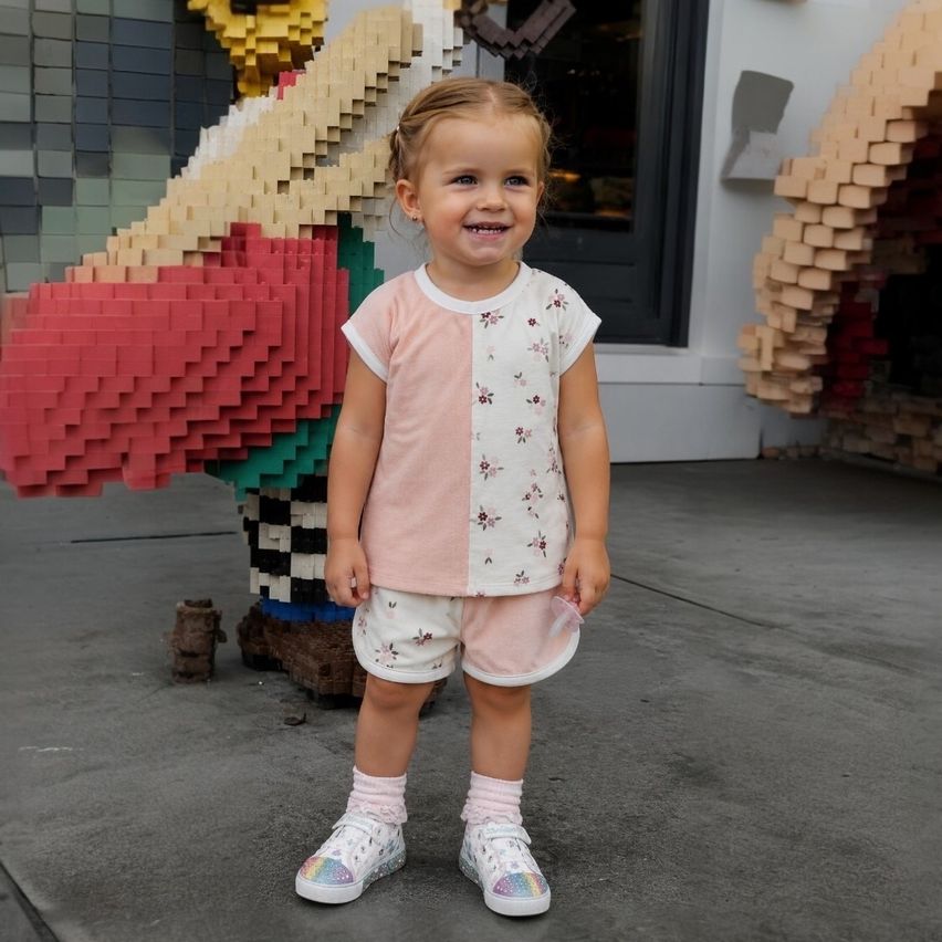Ein kleines Mädchen steht lächelnd vor einer großen Lego-Figur und trägt eine pinke Shorts, ein pink-weißes Oberteil und bunte Turnschuhe.
