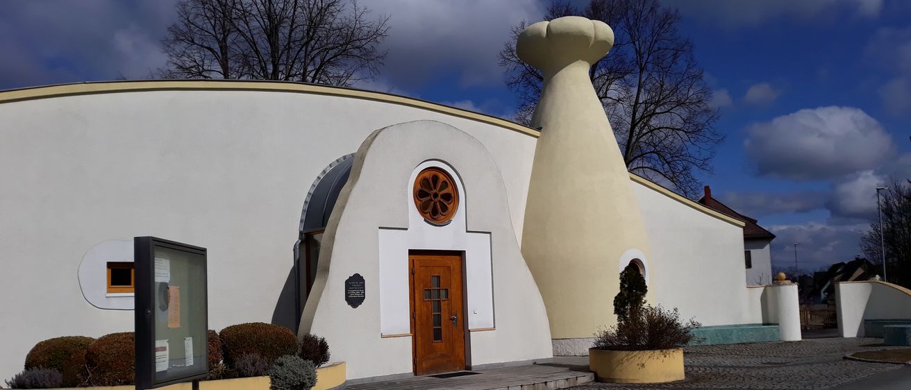 Ein modernes architektonisches Gebäude mit weißer Außenseite, einer Holztür und einem blumenförmigen Fenster darüber. Es hat ein einzigartiges Design mit einem kegelförmigen Turm.