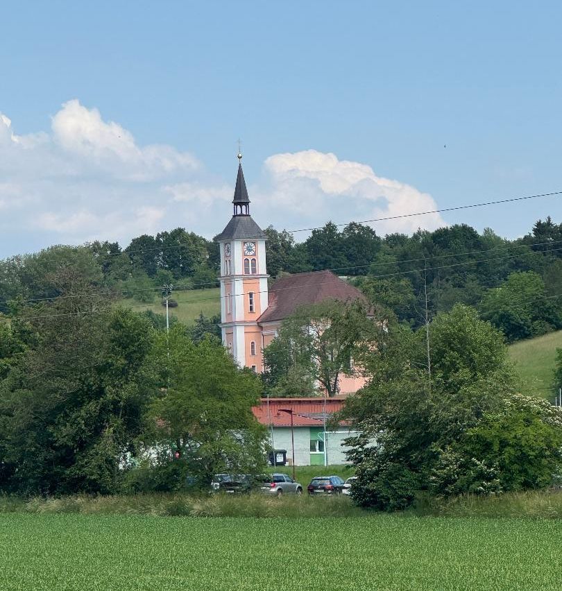 Ein Kirchturm mit einer Uhr ist vor einem Hintergrund von satten grünen Feldern und Bäumen. Der Himmel ist klar mit verstreuten Wolken. Autos sind in der Nähe des Gebäudes geparkt.