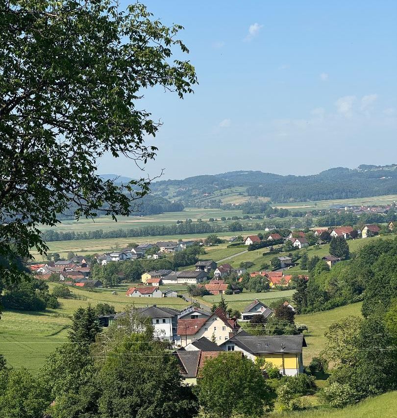 Ein ländliches Dorf mit vielen Häusern umgeben von üppigen grünen Feldern und Bäumen unter einem klaren blauen Himmel mit einigen Wolken.