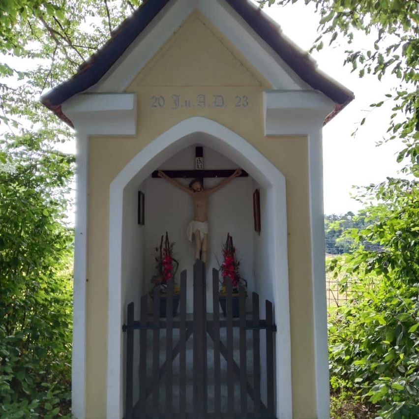 Eine kleine Kapelle mit einem Kruzifix und Blumen davor, datiert auf den 20. Juli 2023. Die Kapelle ist von Grün umgeben.