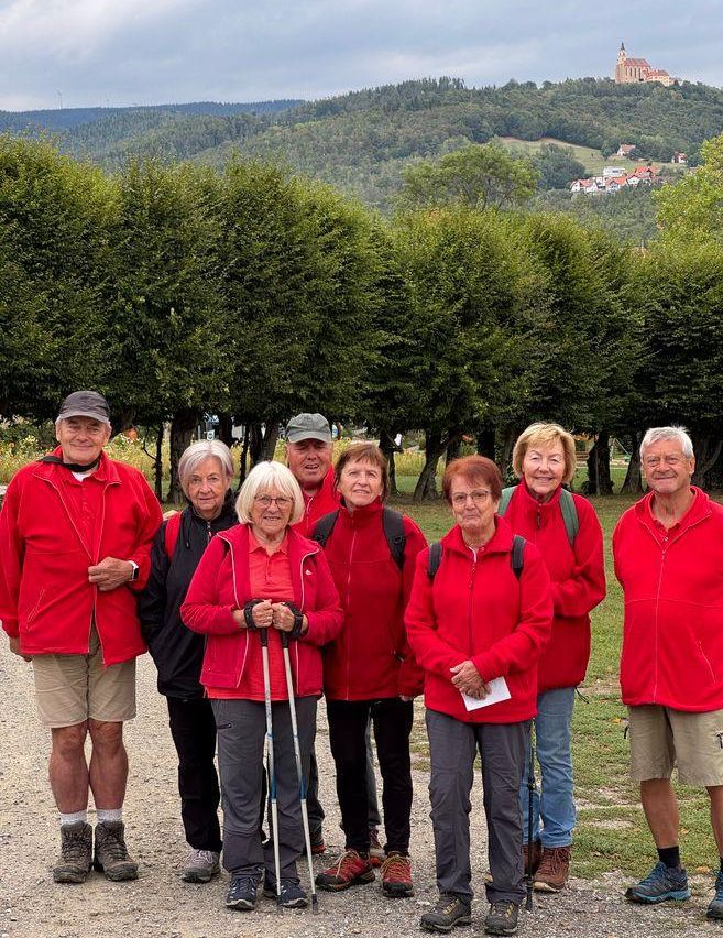 Eine Gruppe älterer Personen in roten Jacken und mit Wanderstöcken steht in einer Reihe auf einem Kiesweg mit Bäumen und einer Burg im Hintergrund.