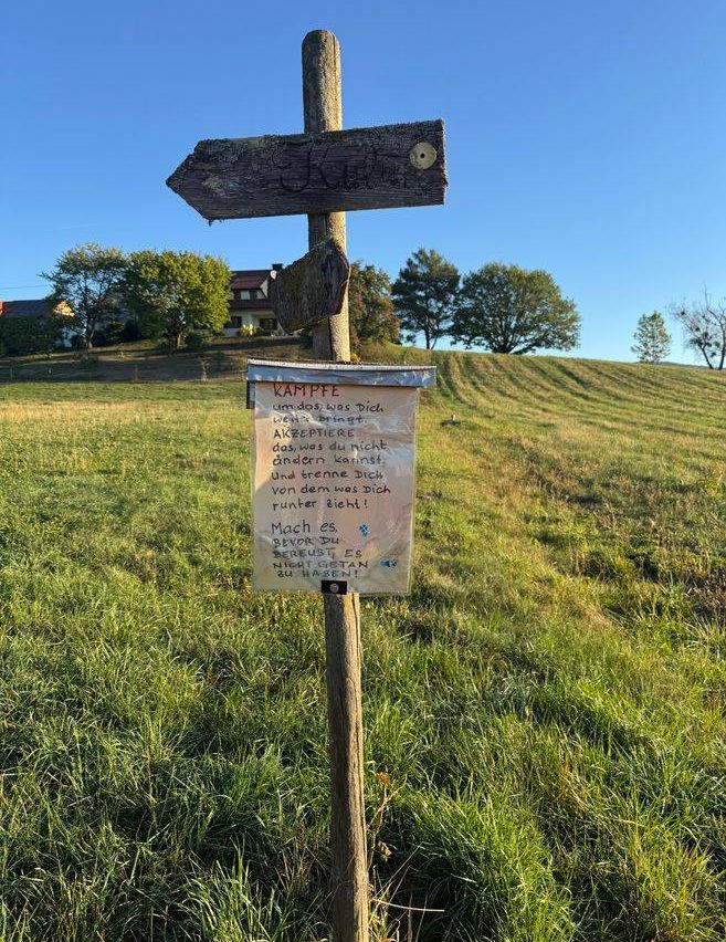 Ein Holzschild mit einer handgeschriebenen Nachricht ist an einem Holzpfosten in einem Grasfeld befestigt.