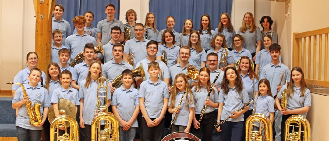 Eine Gruppe junger Musiker, sowohl männlich als auch weiblich, in hellblauen Shirts, posiert für ein Foto mit verschiedenen Instrumenten vor einem blauen Vorhang.