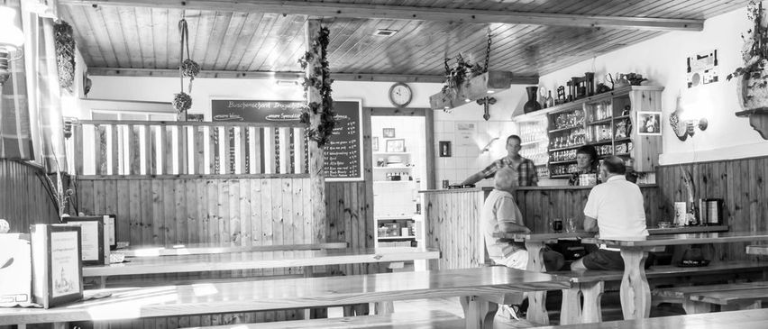 Ein Schwarz-Weiß-Foto eines gemütlichen Restaurants mit Holztischen, Bänken und einer Theke. Ein Menübrett hängt an der Wand, und ein Mann steht hinter der Theke.