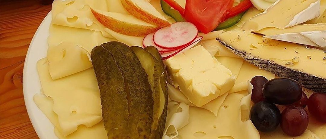 Ein weißer Teller mit verschiedenen Käsescheiben, Gurken, Trauben, einem geschnittenen Ei und einer Mischung aus Gemüse, darunter geschnittene Paprika, Tomaten und Radieschen.