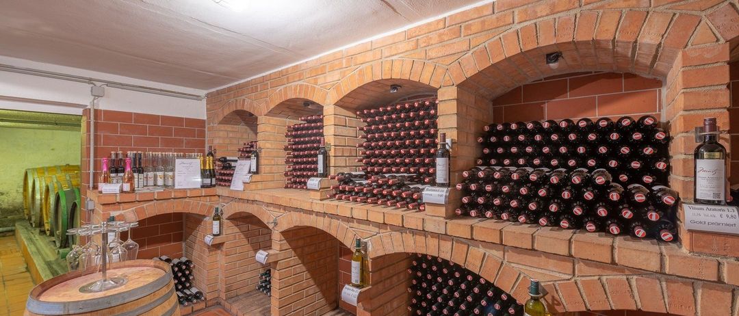 Ein Weinkeller mit gewölbten Ziegelwänden enthält Reihen von Weinflaschen in verschiedenen Farben und Größen. Ein Fass befindet sich links und eine Flasche mit dem Etikett 'Weinkellerung 2017' rechts.