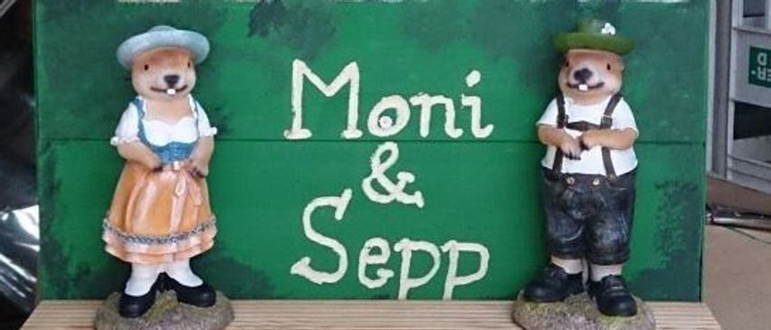 Zwei Keramikfiguren, eine Frau in traditioneller Kleidung und ein Mann in bayrischer Tracht, stehen vor einem grünen Schild mit den Namen 'Moni & Sepp' in weißer Schrift.