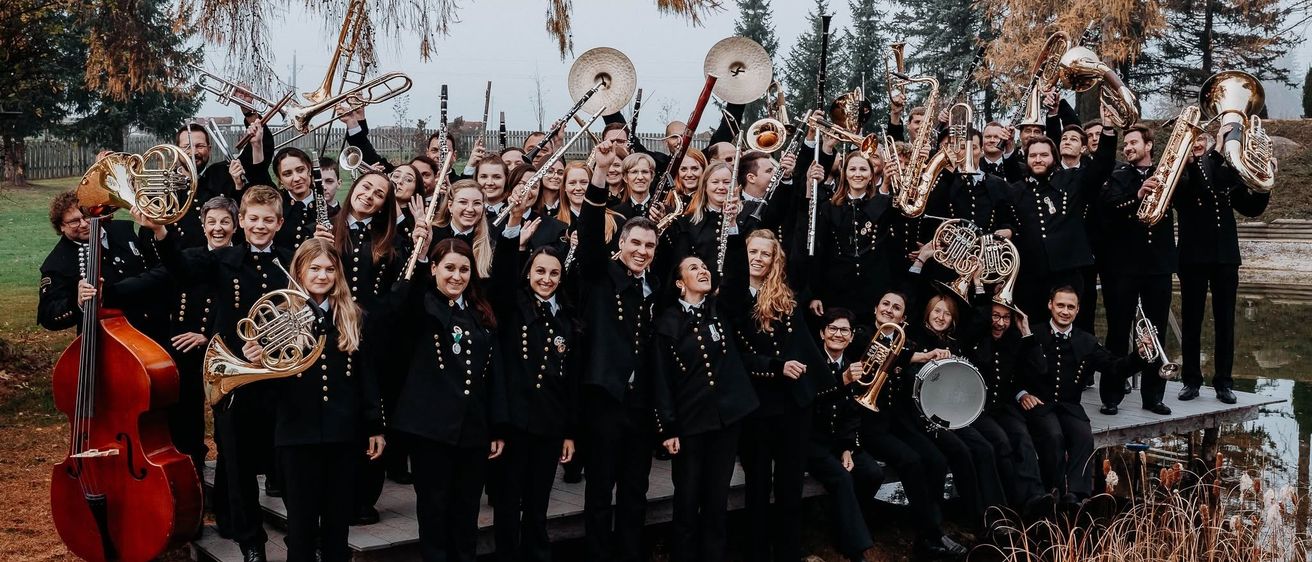 Eine Gruppe uniformierter Musiker posiert für ein Foto im Freien mit Instrumenten. Der Hintergrund ist ein grüner Rasen mit Bäumen.