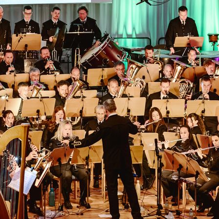 Ein Orchester spielt auf der Bühne mit verschiedenen Instrumenten. Ein Dirigent leitet die Musiker. Ein großer Bildschirm ist im Hintergrund, und grüne Vorhänge sind auf beiden Seiten.