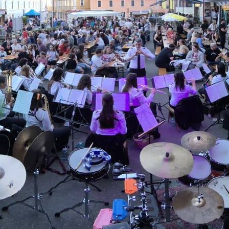 Ein großes Ensemble spielt auf der Straße im Freien. Eine Menge schaut zu. Einige sitzen an Tischen mit Essen, während andere stehen. Die Band besteht aus Blasinstrumenten, Trommeln und Becken.