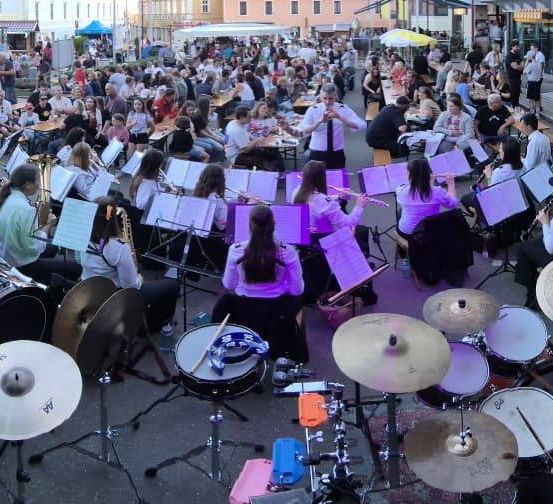 Ein großes Ensemble spielt auf der Straße im Freien. Eine Menge schaut zu. Einige sitzen an Tischen mit Essen, während andere stehen. Die Band besteht aus Blasinstrumenten, Trommeln und Becken.