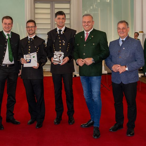 Eine Gruppe von Menschen, darunter Männer und Frauen, stehen auf einem roten Teppich in einem formellen Rahmen. Sie sind alle in Anzügen und Krawatten gekleidet. Sie posieren für ein Foto.