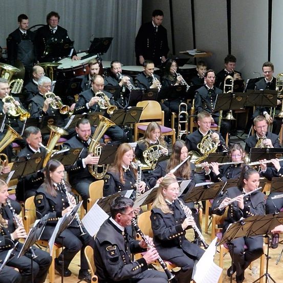 Ein Militärorchester in Uniform spielt verschiedene Instrumente, darunter Trompeten, Posaunen, Flöten und Klarinetten, unter der Leitung eines Dirigenten auf der Bühne.